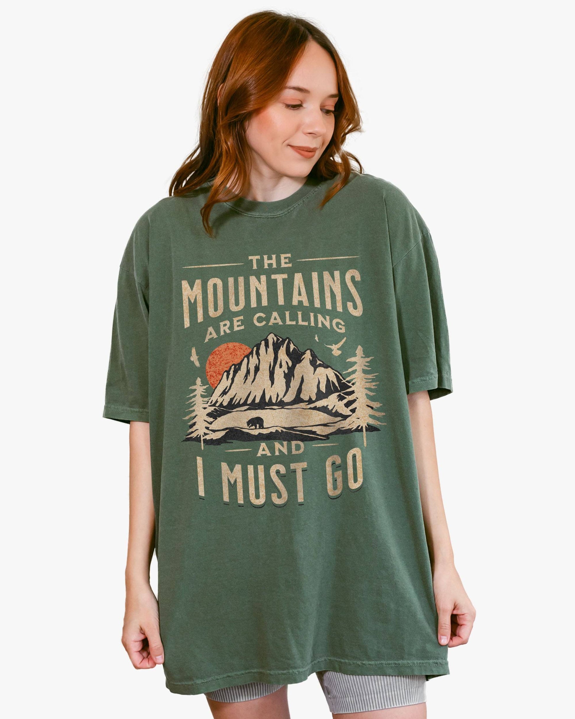Woman wearing moss shirt with phrase ‘The Mountains Are Calling And I Must Go’ and retro mountain nature graphic.