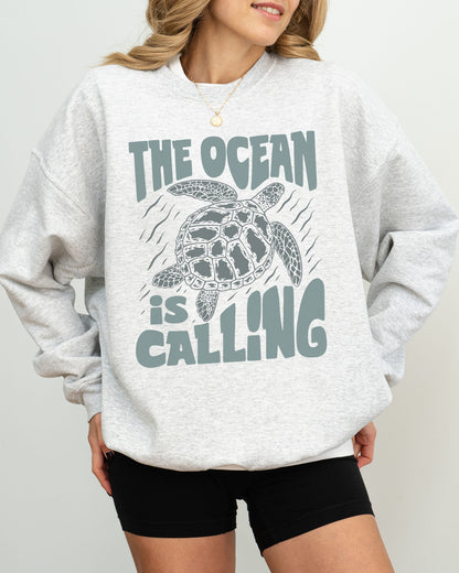 Model wearing ash color sweatshirt with 'The Ocean Is Calling' graphic design