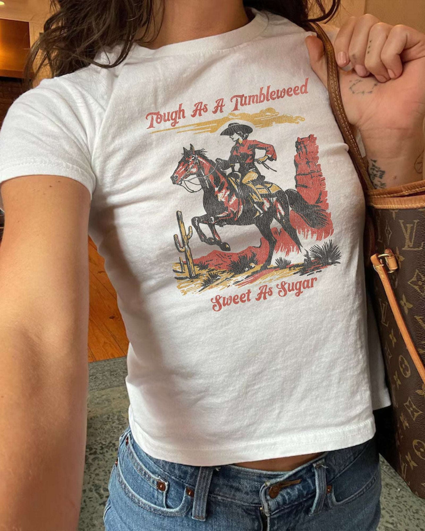Woman wearing white baby tee with retro country western graphic and text Tough As A Tumbleweed Sweet As Sugar