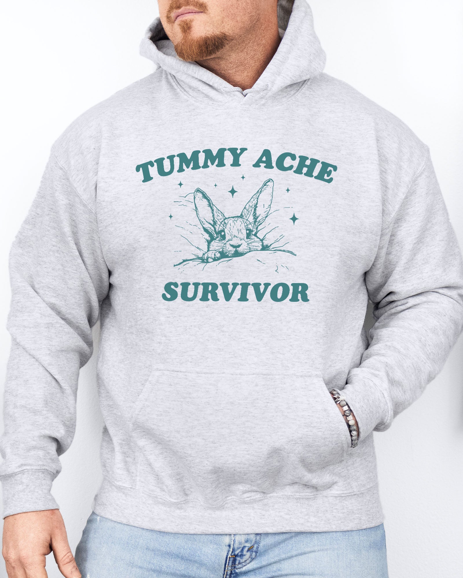 Man wearing an ash gray hoodie with a funny retro-style graphic of a bunny lying in bed and the quote 'Tummy Ache Survivor'.