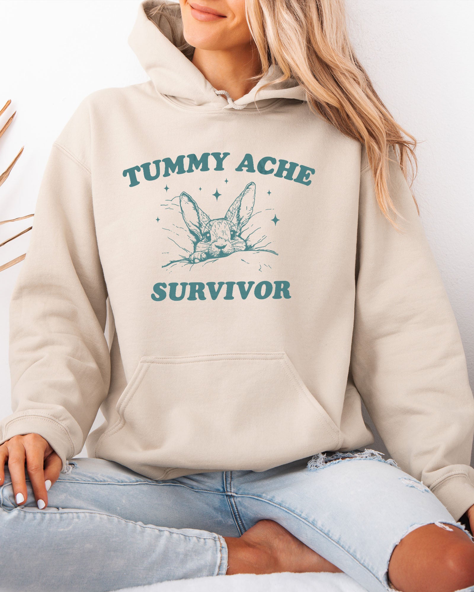 Woman wearing a sand beige hoodie with a funny retro-style graphic of a bunny lying in bed and the quote 'Tummy Ache Survivor'.
