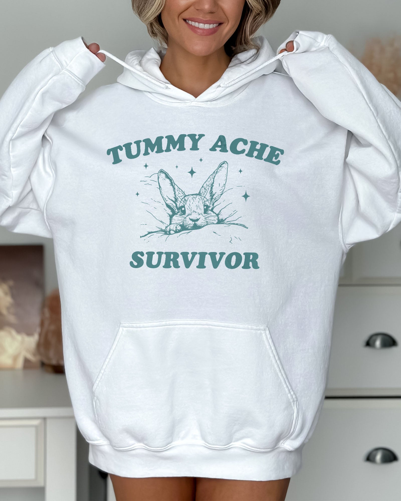 Woman wearing a white hoodie with a funny retro-style graphic of a bunny lying in bed and the quote 'Tummy Ache Survivor'.