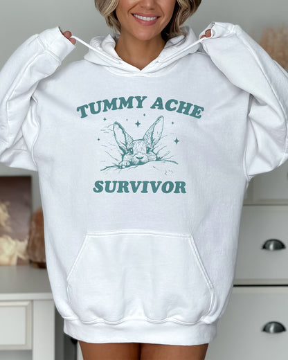 Woman wearing a white hoodie with a funny retro-style graphic of a bunny lying in bed and the quote 'Tummy Ache Survivor'.