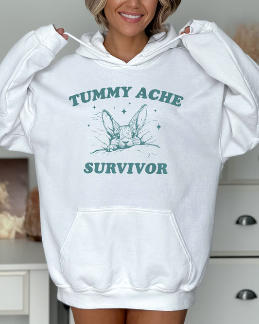 Woman wearing a white hoodie with a funny retro-style graphic of a bunny lying in bed and the quote 'Tummy Ache Survivor'.