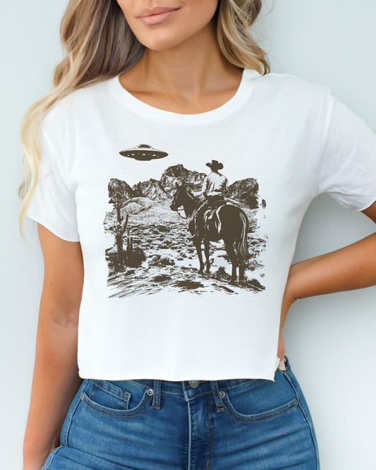 Woman wearing white crop top featuring a UFO and western desert scenery graphic.