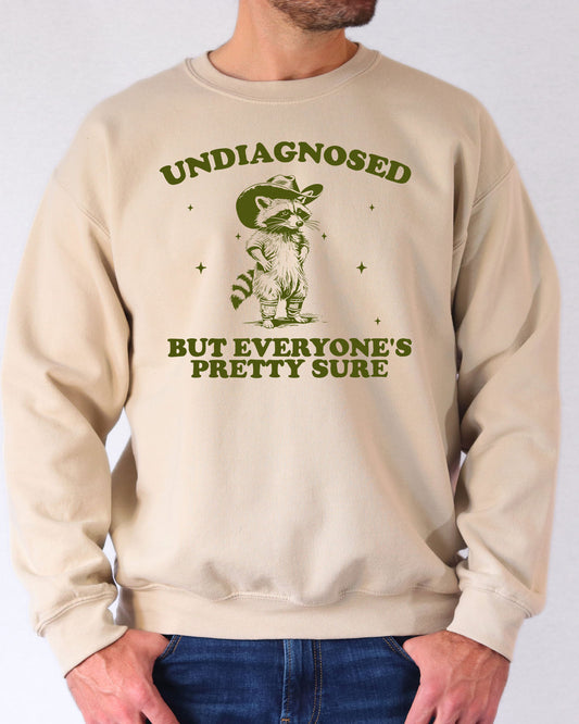 Man wearing a sand beige sweatshirt featuring a funny retro-style cowboy raccoon graphic and the quote 'Undiagnosed But Everyone Is Pretty Sure'.