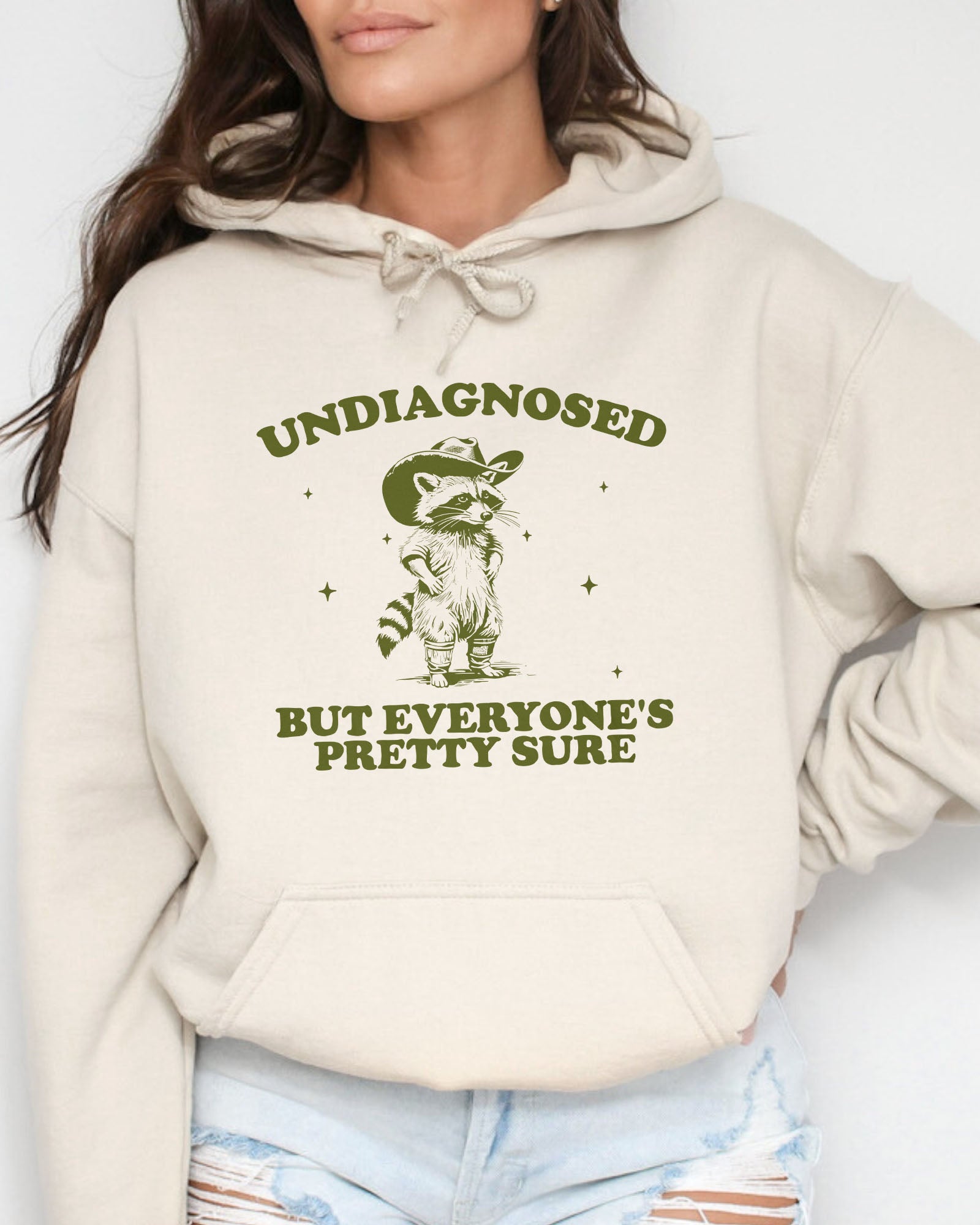 Woman wearing sand beige hoodie with a funny retro cowboy raccoon graphic and the quote 'Undiagnosed, But Everyone's Pretty Sure'.
