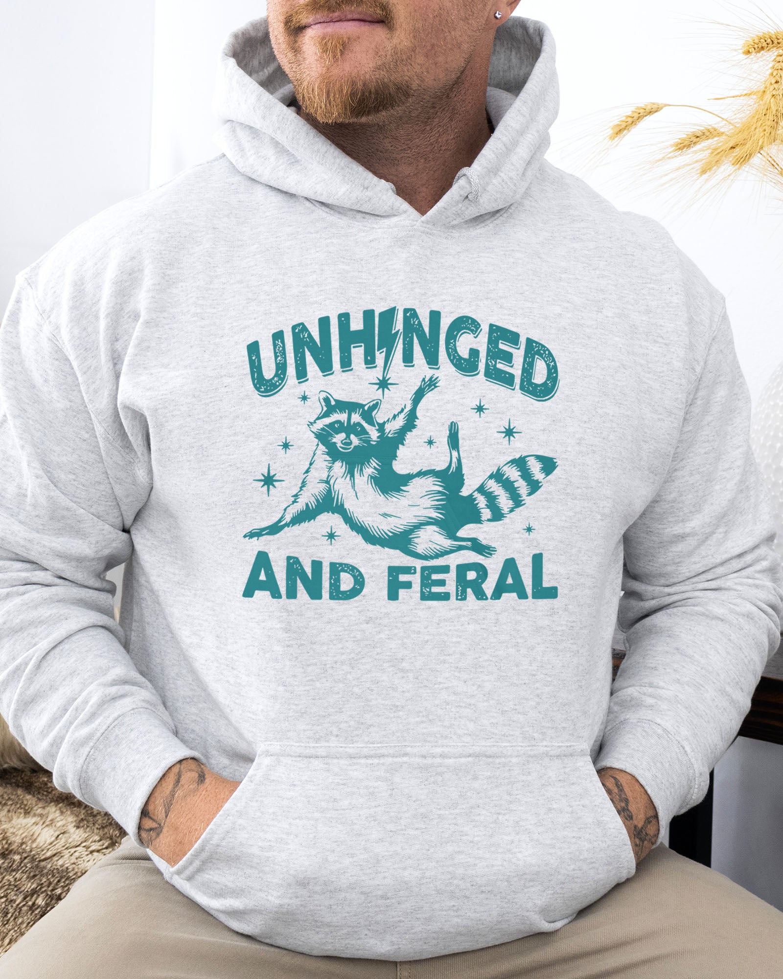 Man wearing an ash-colored hoodie featuring a funny retro raccoon graphic and the quote 'Unhinged And Feral'.