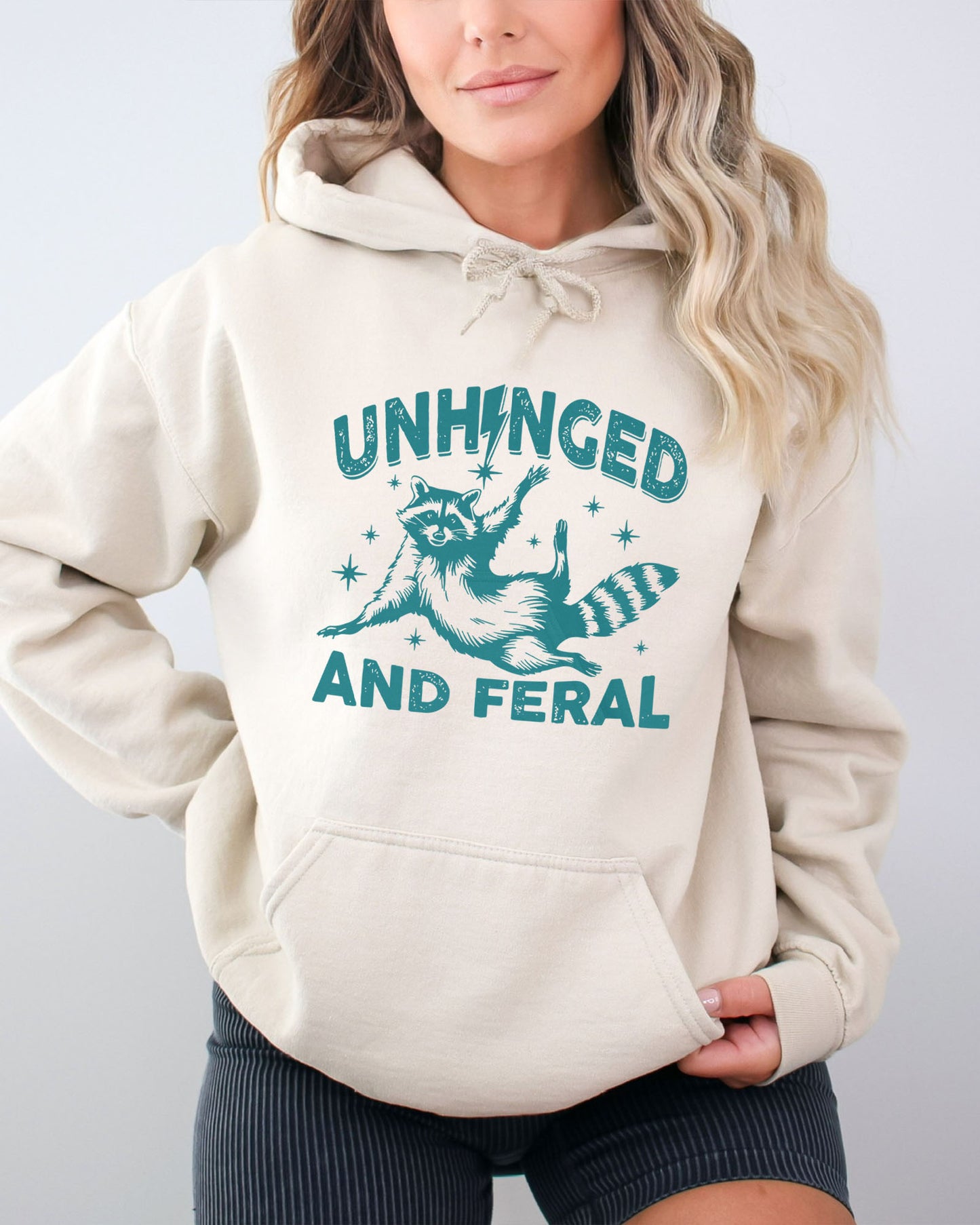 Woman wearing a sand-colored hoodie featuring a funny retro raccoon graphic and the quote 'Unhinged And Feral'.