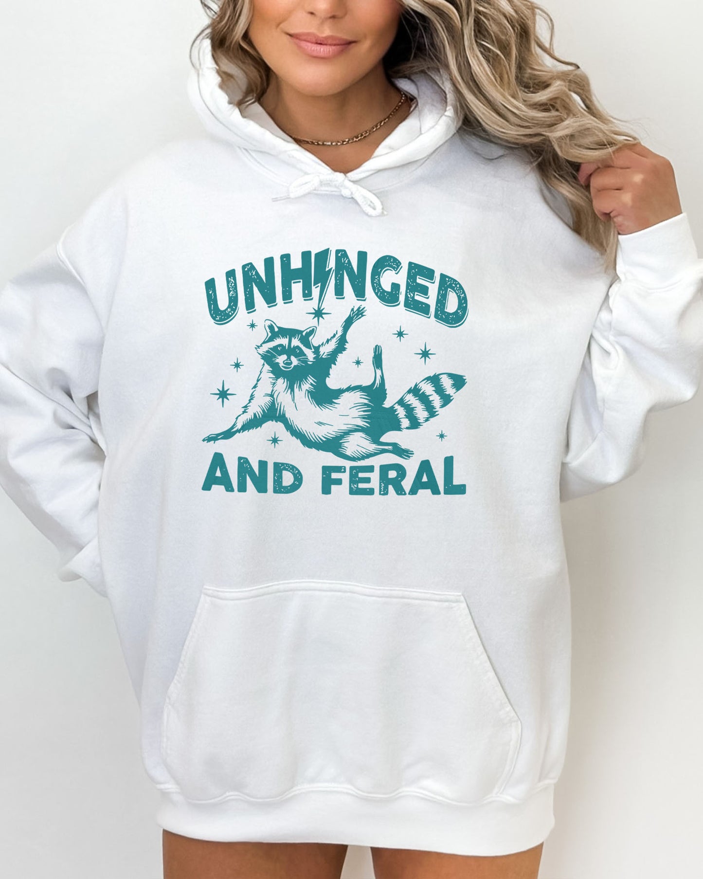 Woman wearing a white-colored hoodie featuring a funny retro raccoon graphic and the quote 'Unhinged And Feral'.