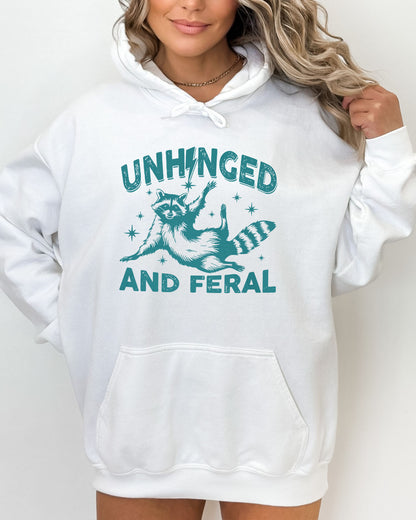 Woman wearing a white-colored hoodie featuring a funny retro raccoon graphic and the quote 'Unhinged And Feral'.