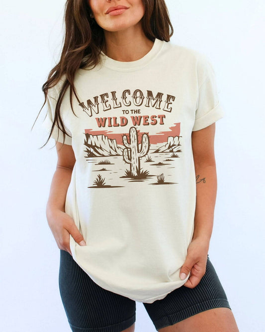 Woman wearing an ivory 'Welcome To The Wild West' shirt with a retro western graphic of a desert and cactus.