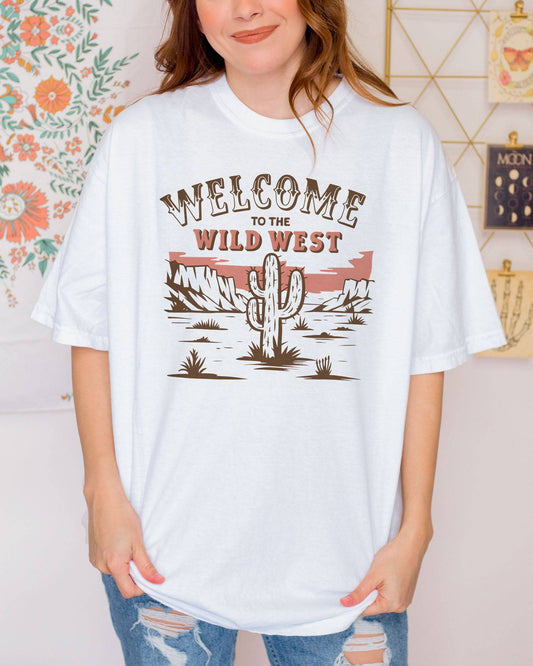 Woman wearing a white 'Welcome To The Wild West' shirt with a retro western graphic of a desert and cactus.