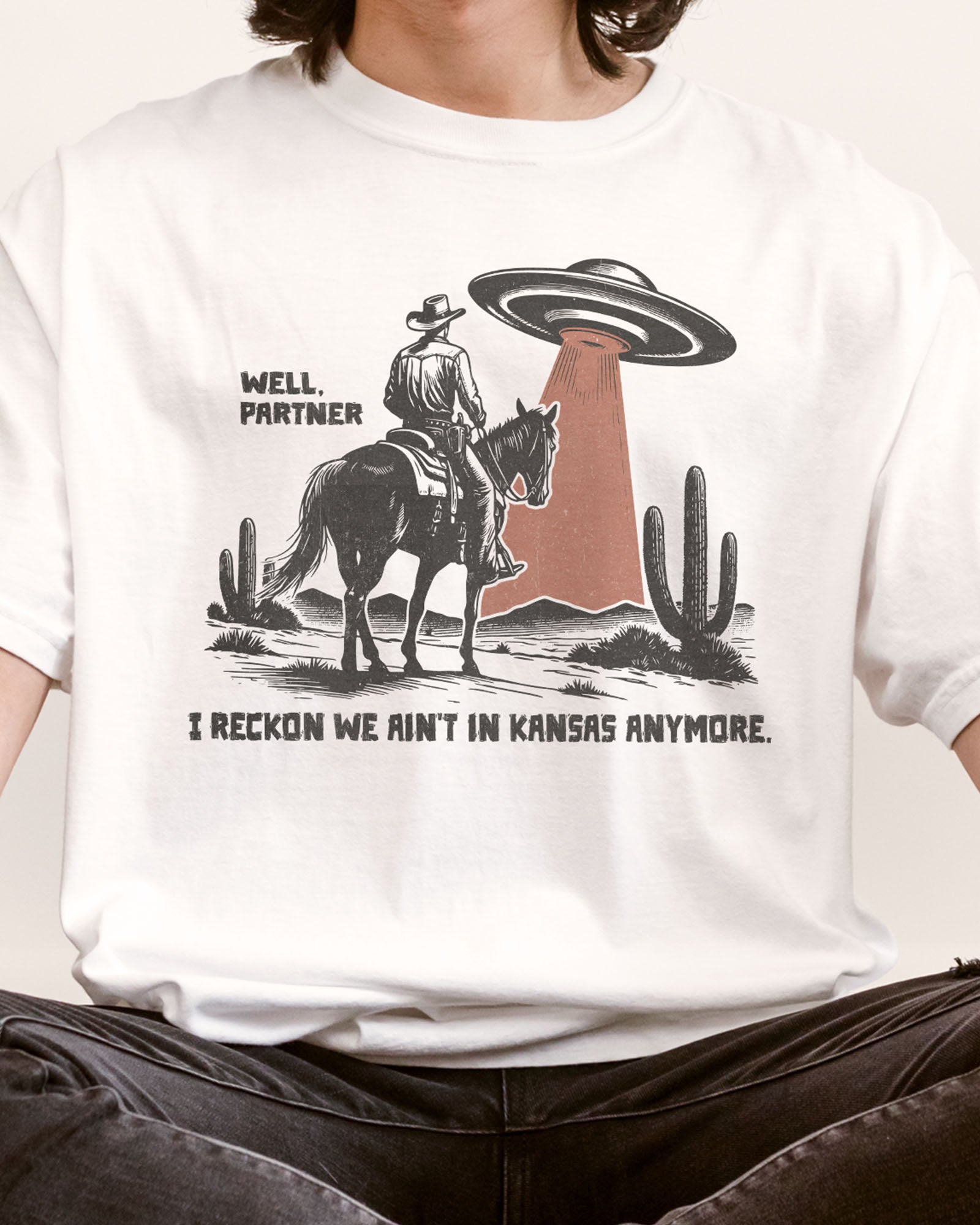 Close-up of "Well Partner I Reckon We Ain't In Kansas Anymore" shirt and a funny retro graphic of a cowboy riding a horse through a desert with an UFO appearing.