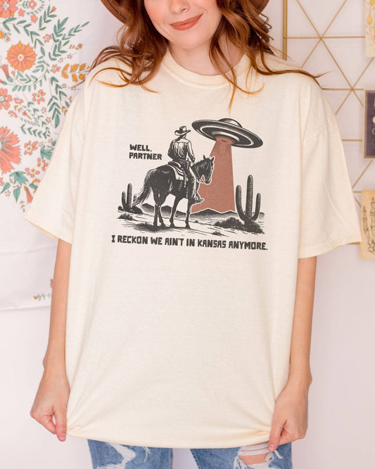 Woman wearing ivory color shirt with quote "Well Partner I Reckon We Ain't In Kansas Anymore" and a funny retro graphic of a cowboy riding a horse through a desert with an UFO appearing.