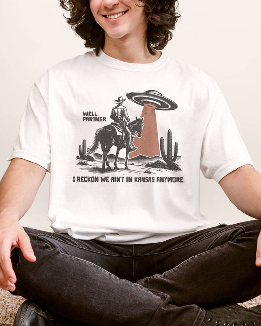 Man wearing white shirt with quote "Well Partner I Reckon We Ain't In Kansas Anymore" and a funny retro graphic of a cowboy riding a horse through a desert with an UFO appearing.
