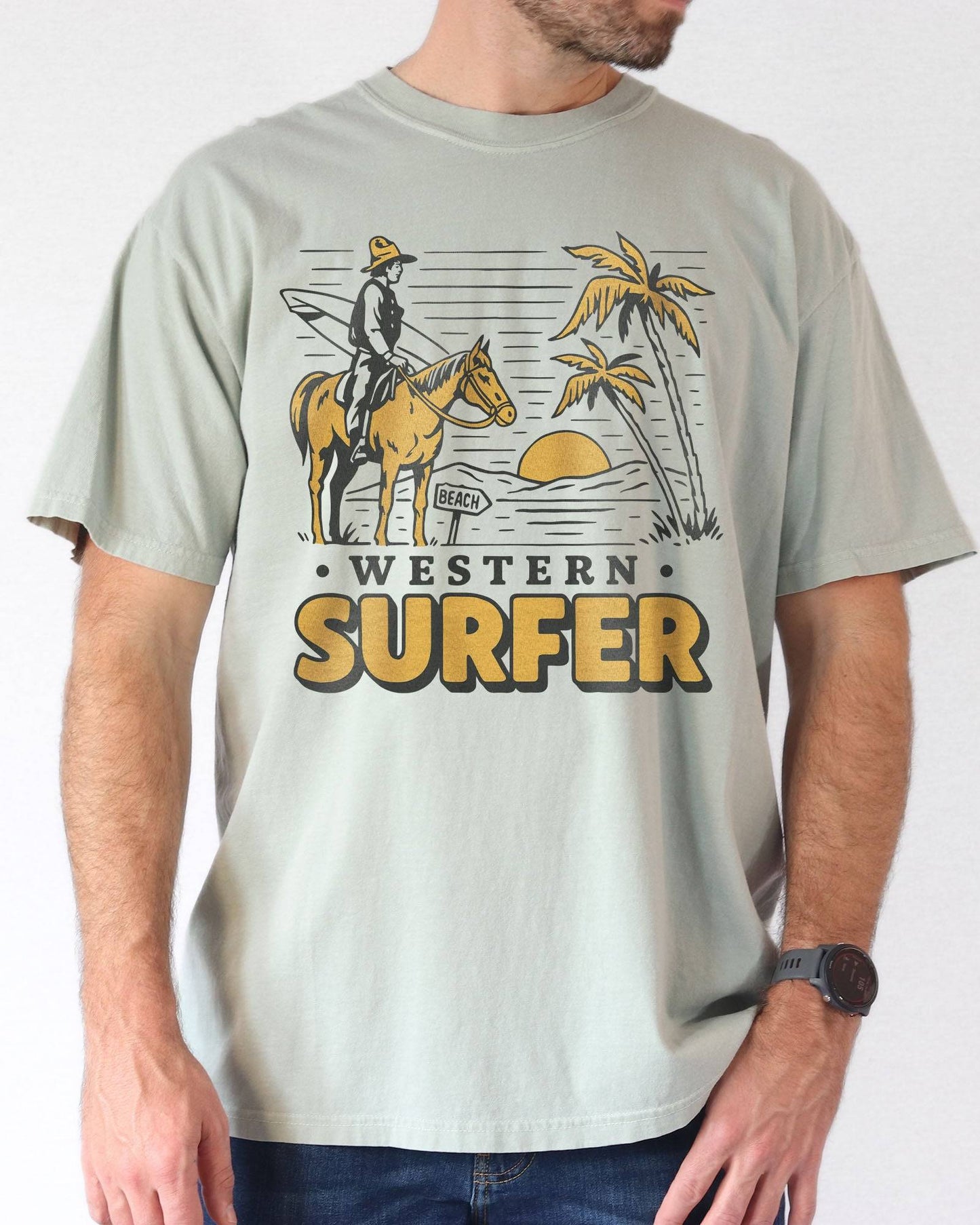 Man wearing a bay-colored 'Western Surfer' shirt with a retro graphic of a cowboy on a horse in a desert, following a beach signpost.