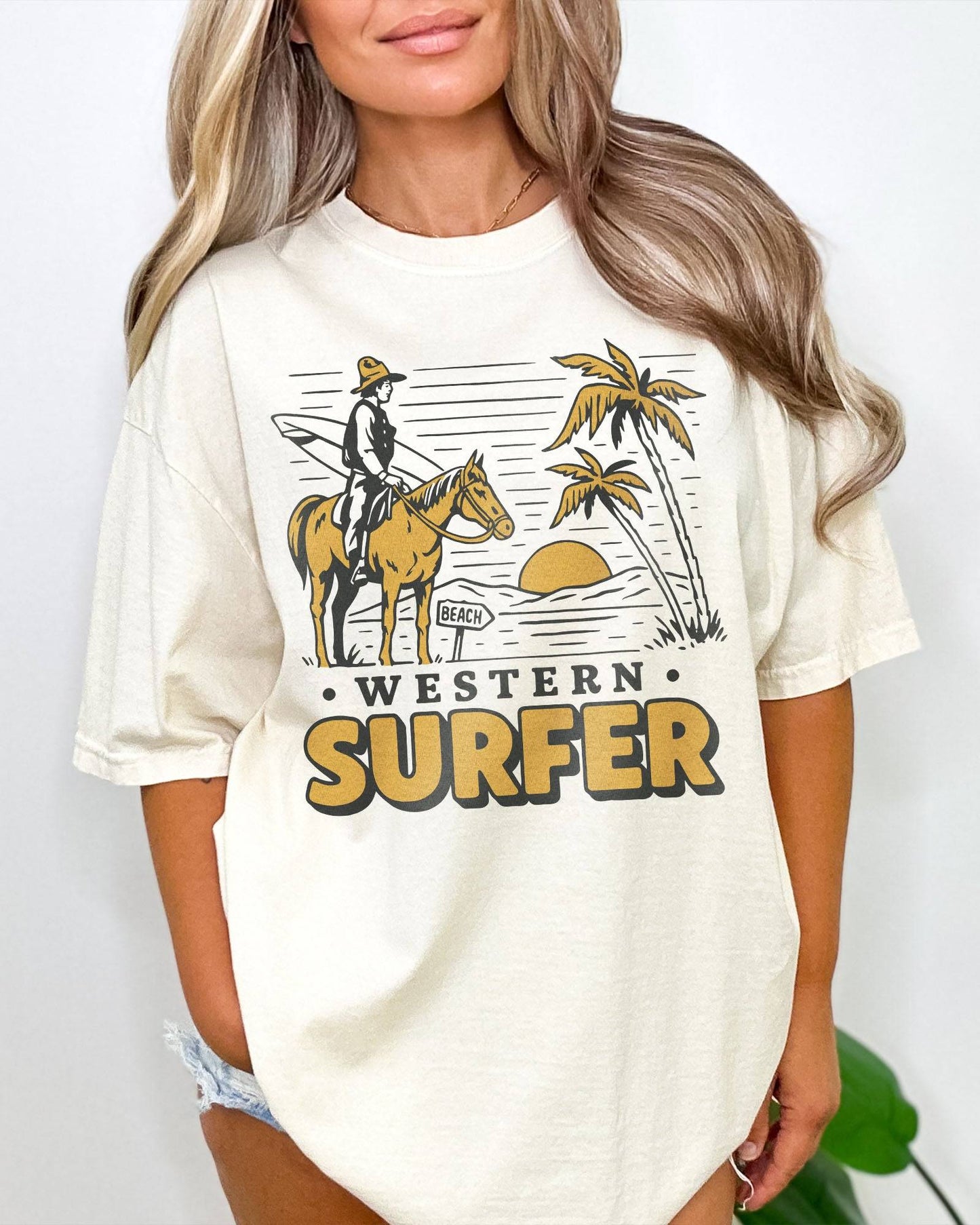 Woman wearing an ivory-colored 'Western Surfer' shirt with a retro graphic of a cowboy on a horse in a desert, following a beach signpost.