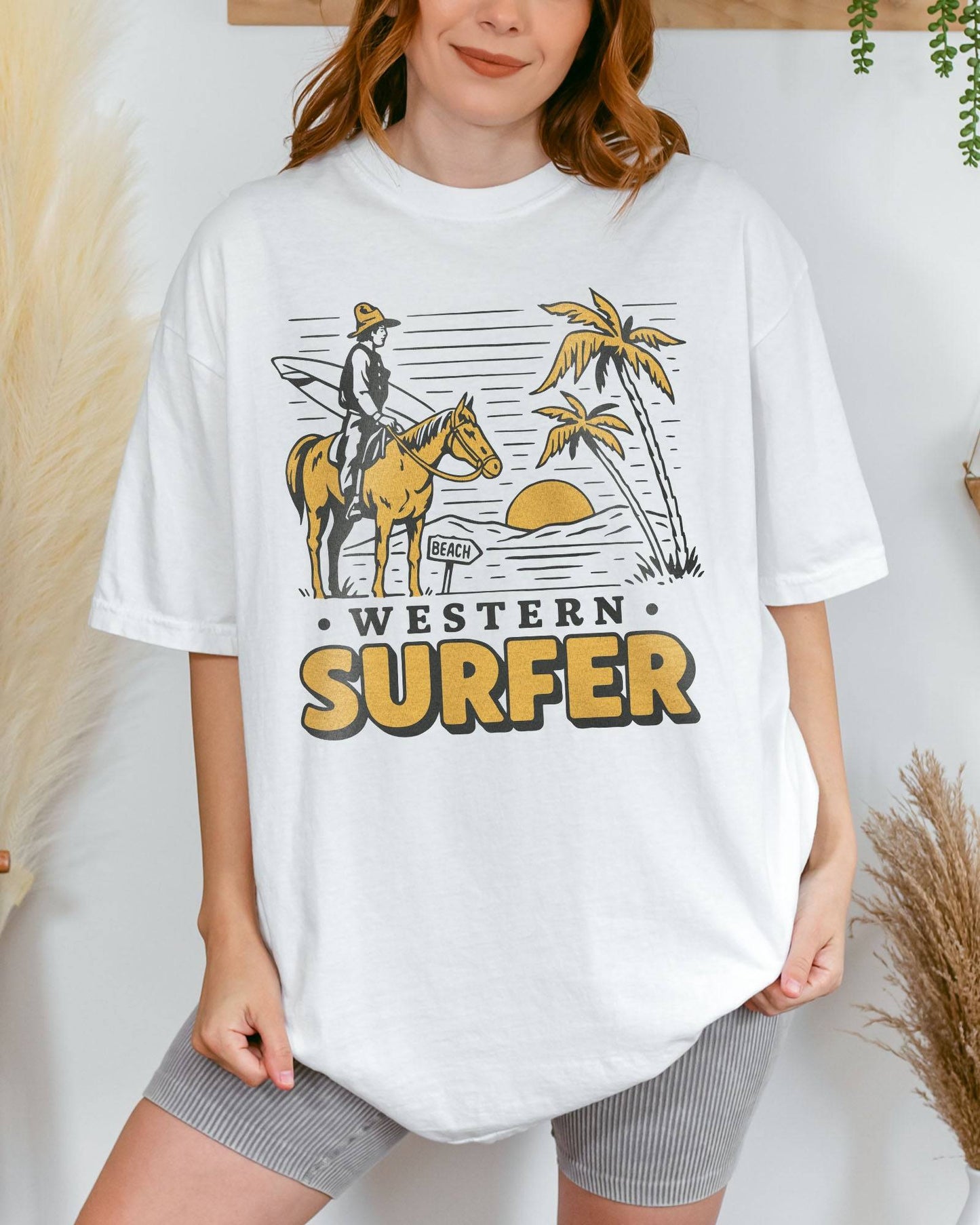 Woman wearing a white 'Western Surfer' shirt with a retro graphic of a cowboy on a horse in a desert, following a beach signpost.