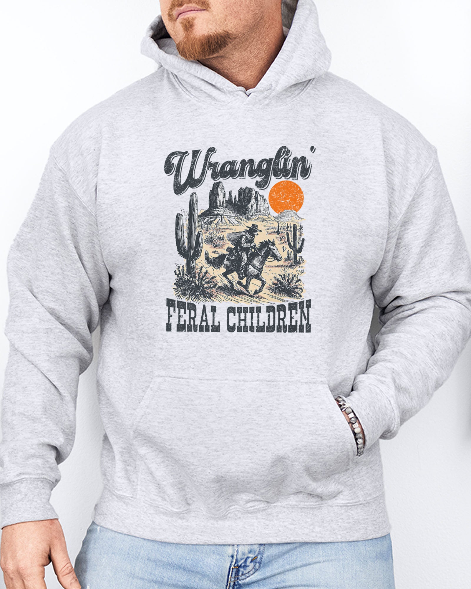 Man wearing an ash-colored hoodie with a retro western graphic of a cowboy raccoon riding a horse and the quote 'Wranglin' Feral Children'.