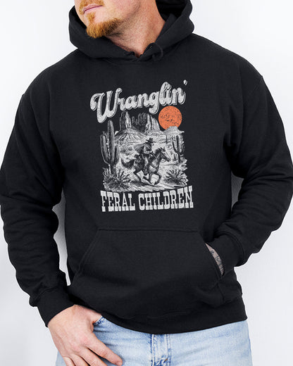 Man wearing a black-colored hoodie with a retro western graphic of a cowboy raccoon riding a horse and the quote 'Wranglin' Feral Children'.