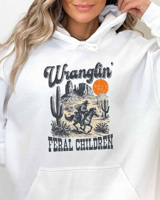 Close up of woman wearing a white-colored hoodie with a retro western graphic of a cowboy raccoon riding a horse and the quote 'Wranglin' Feral Children'.