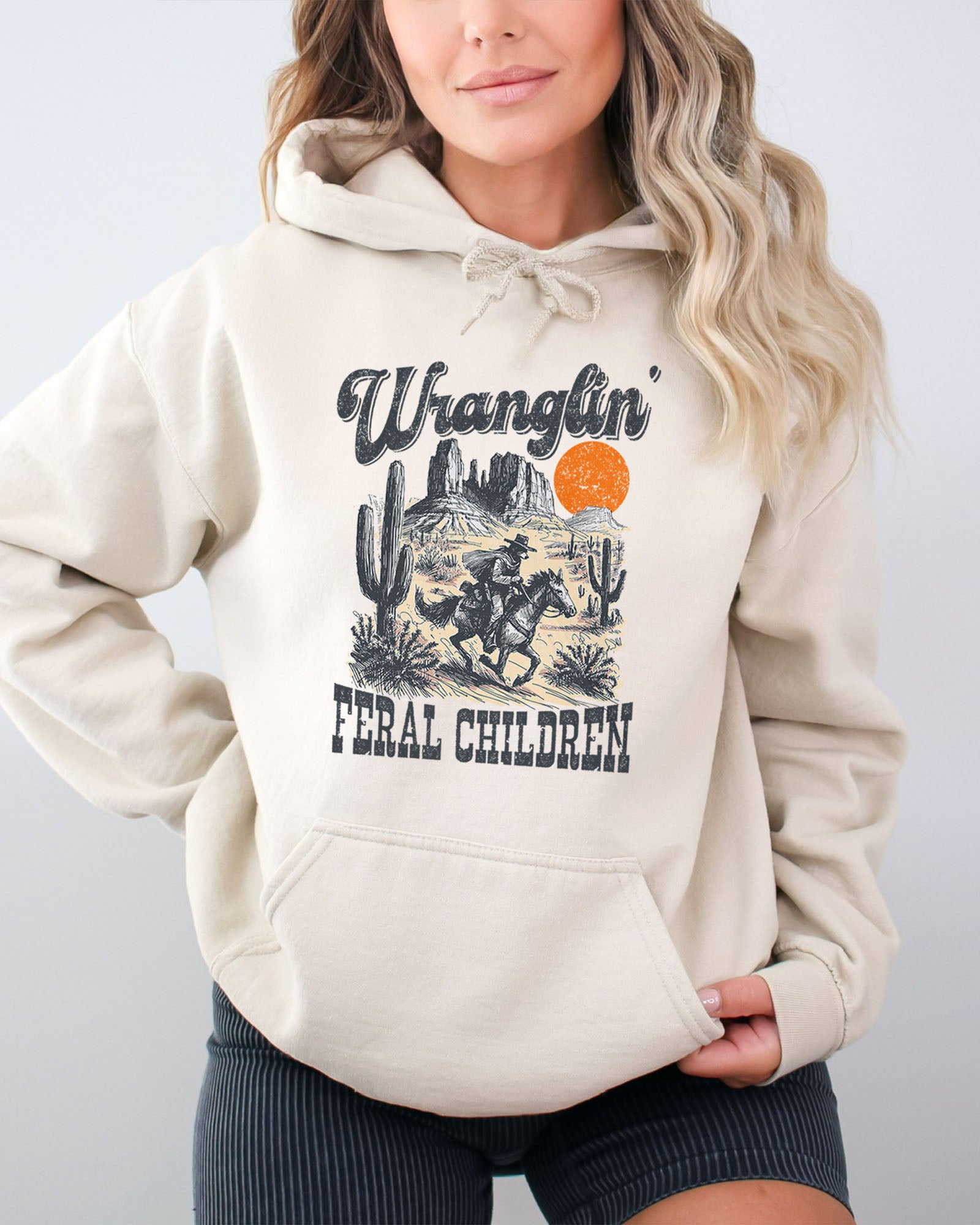 Woman wearing a sand-colored hoodie with a retro western graphic of a cowboy raccoon riding a horse and the quote 'Wranglin' Feral Children'.