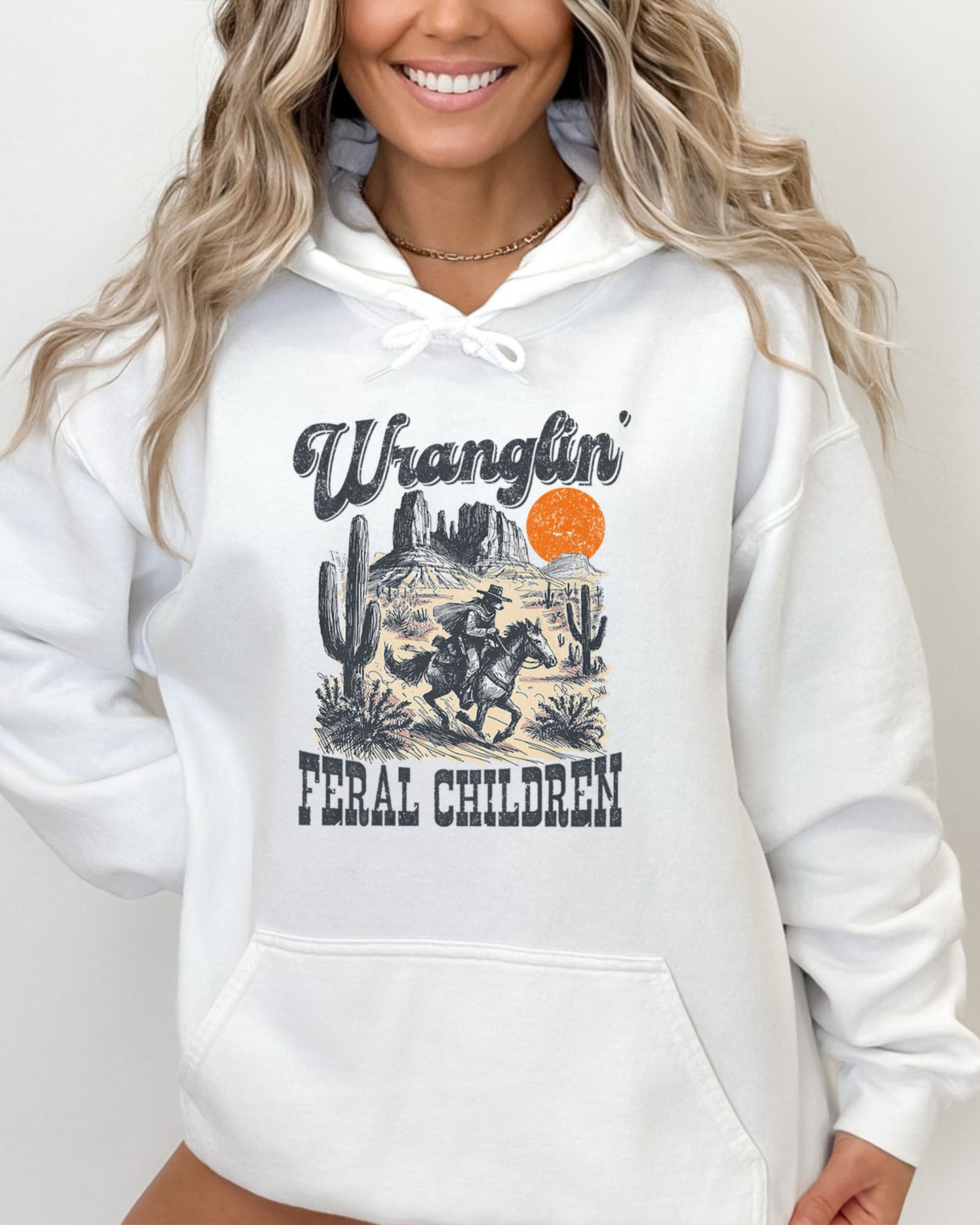 Woman wearing a white-colored hoodie with a retro western graphic of a cowboy raccoon riding a horse and the quote 'Wranglin' Feral Children'.