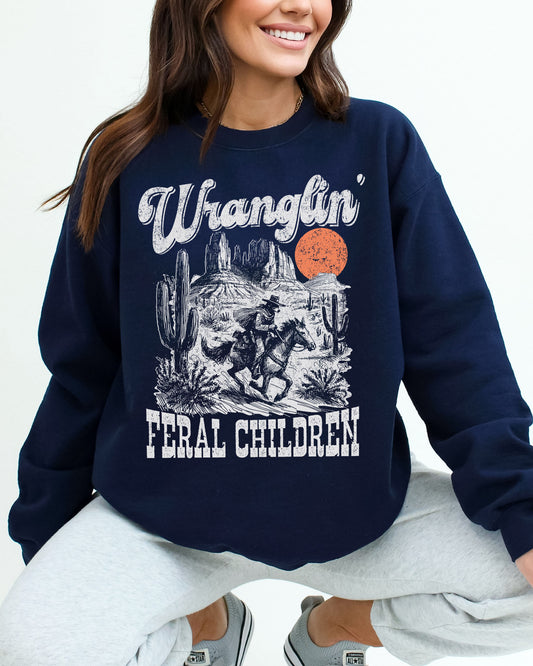 Woman wearing a navy blue sweatshirt featuring a retro graphic of a cowboy riding a horse through a Western desert scene with the quote 'Wranglin' Feral Children'.
