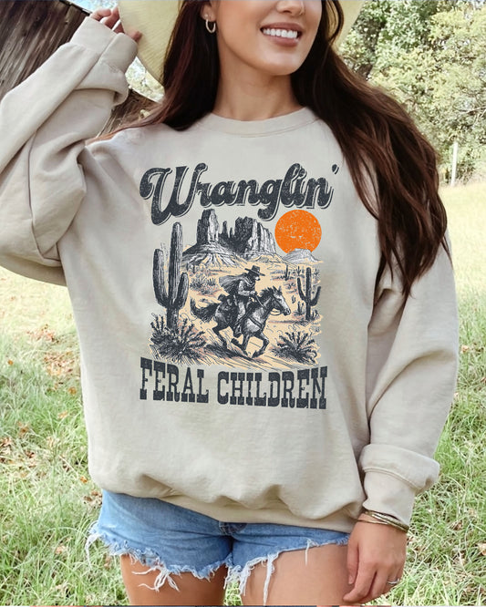 Woman wearing a sand beige sweatshirt featuring a retro graphic of a cowboy riding a horse through a Western desert scene with the quote 'Wranglin' Feral Children'.