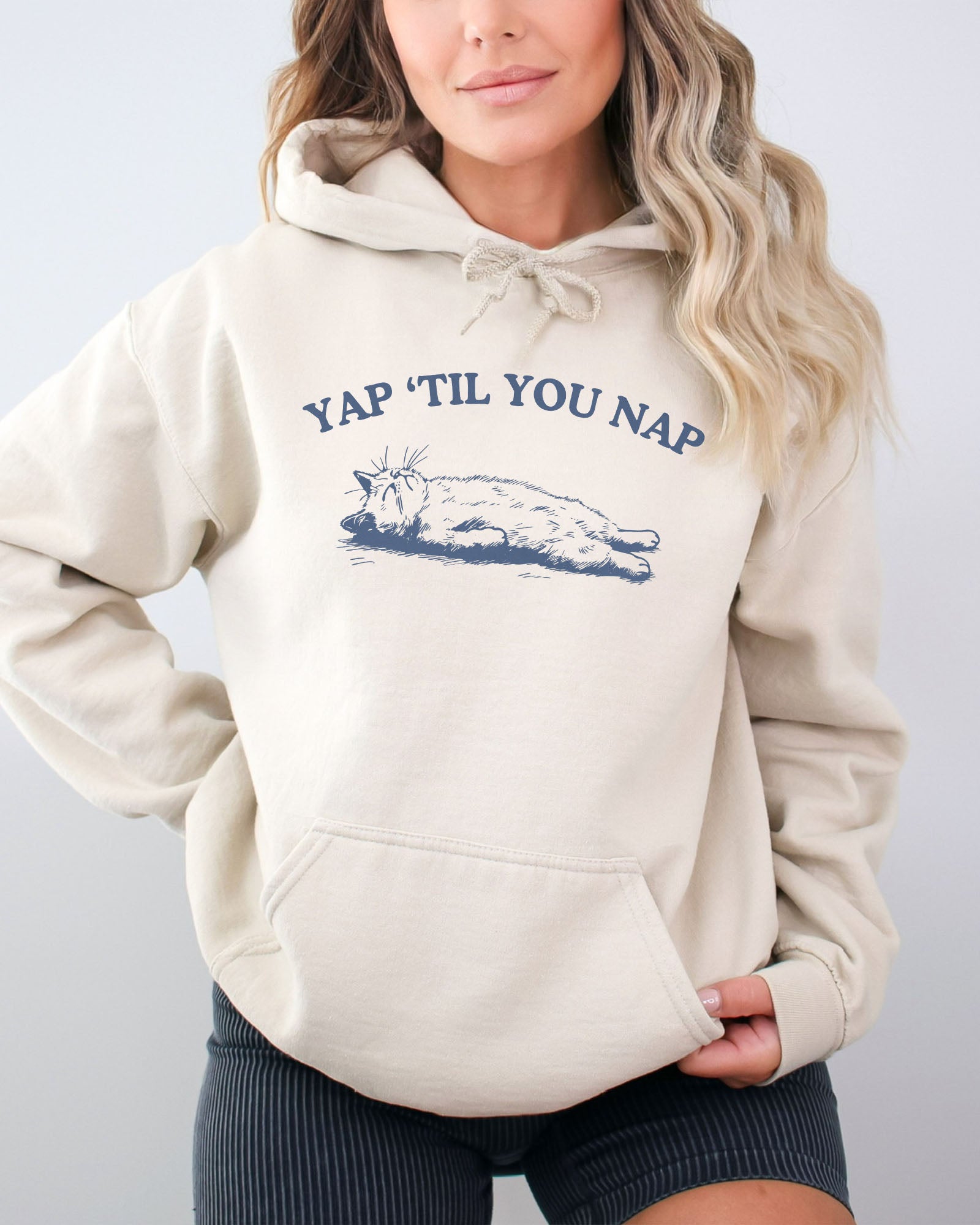 Woman wearing a sand beige hoodie featuring a funny retro sleeping cat graphic and the quote 'Yap 'Til You Nap'.