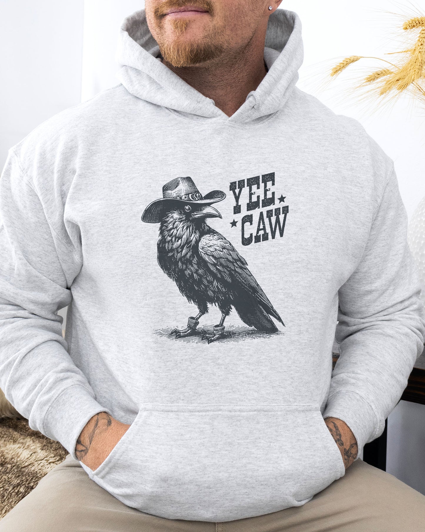 Man wearing an ash-colored hoodie featuring a funny retro cowboy crow graphic and the quote 'Yee Caw'.