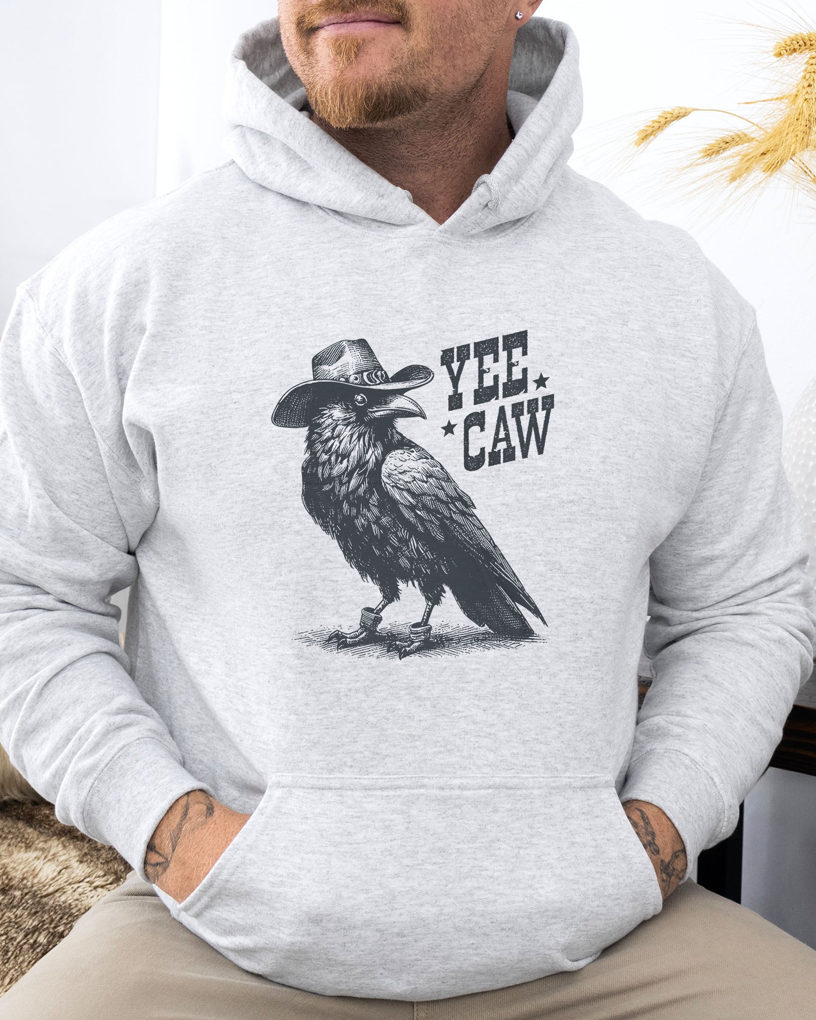 Man wearing an ash-colored hoodie featuring a funny retro cowboy crow graphic and the quote 'Yee Caw'.