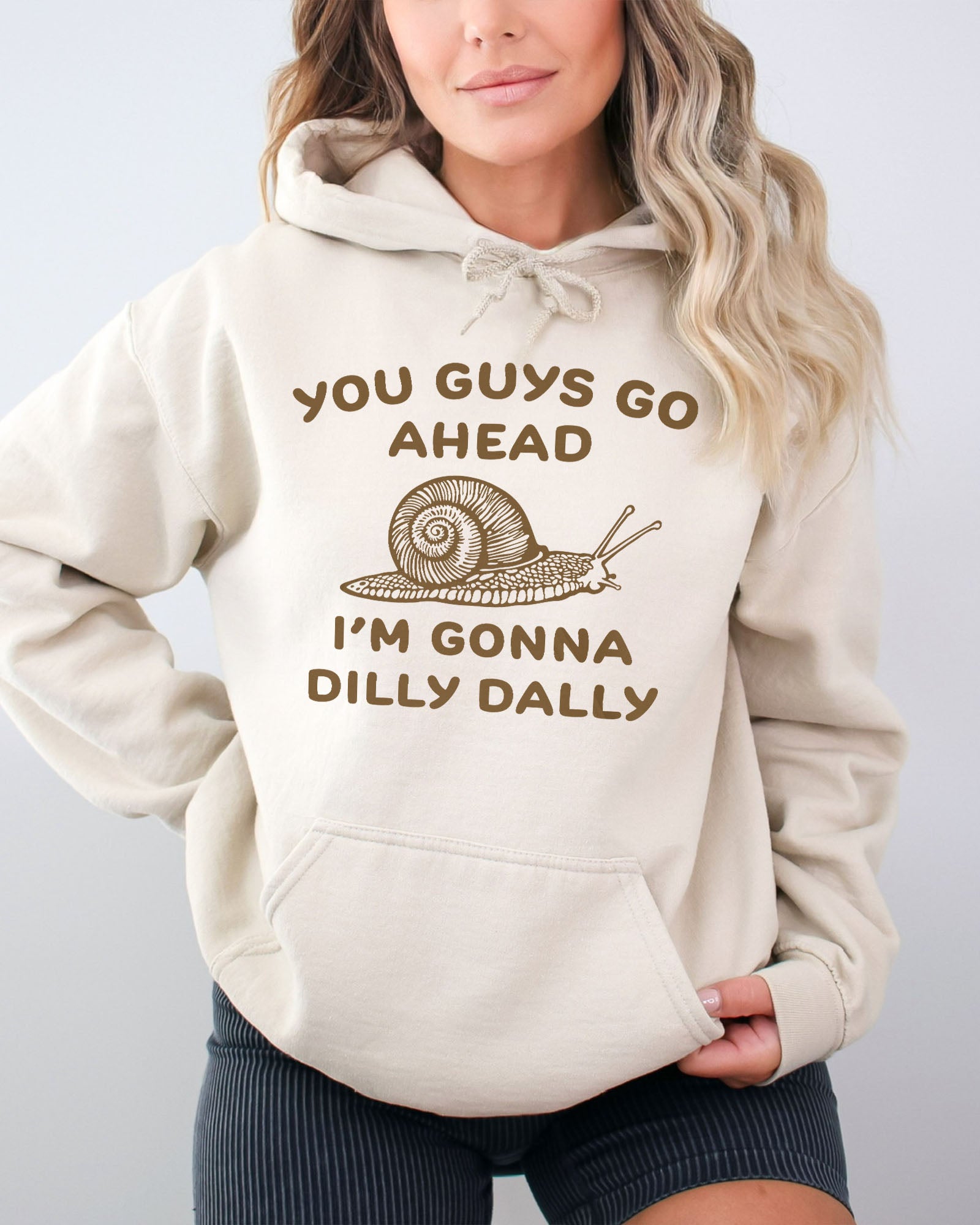Woman wearing a sand beige hoodie with a funny retro-style snail graphic and the quote 'You Guys Go Ahead, I'm Gonna Dilly Dally'.