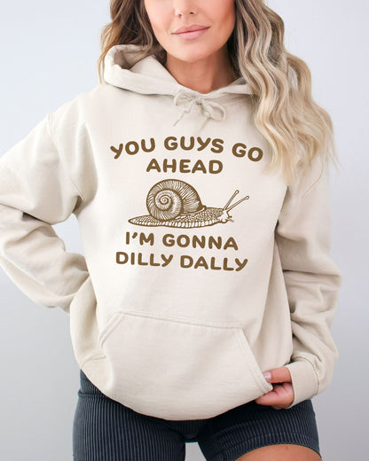 Woman wearing a sand beige hoodie with a funny retro-style snail graphic and the quote 'You Guys Go Ahead, I'm Gonna Dilly Dally'.