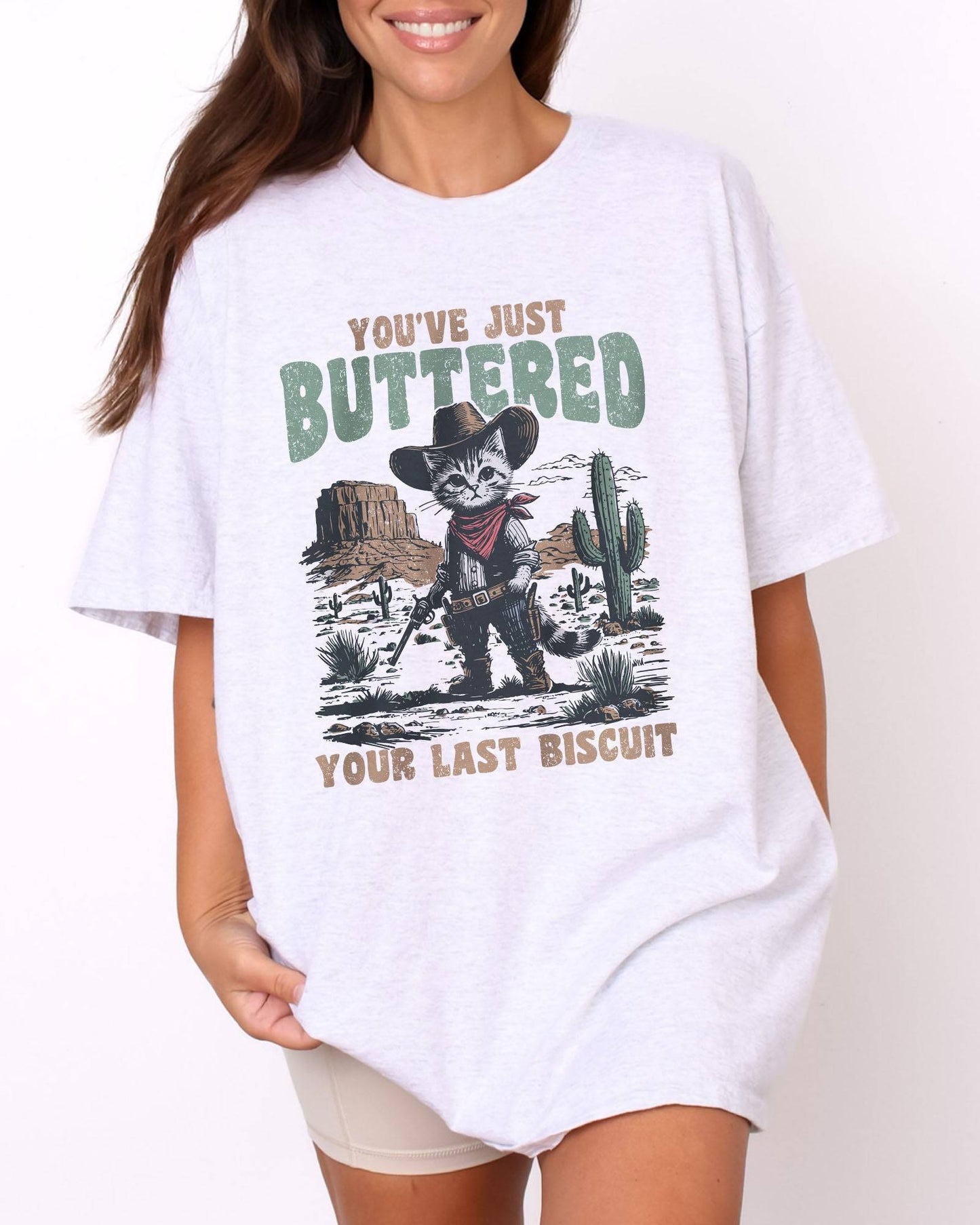Model wearing ash color shirt with 'You've Just Buttered Your Last Biscuit' cowboy kitten graphic design