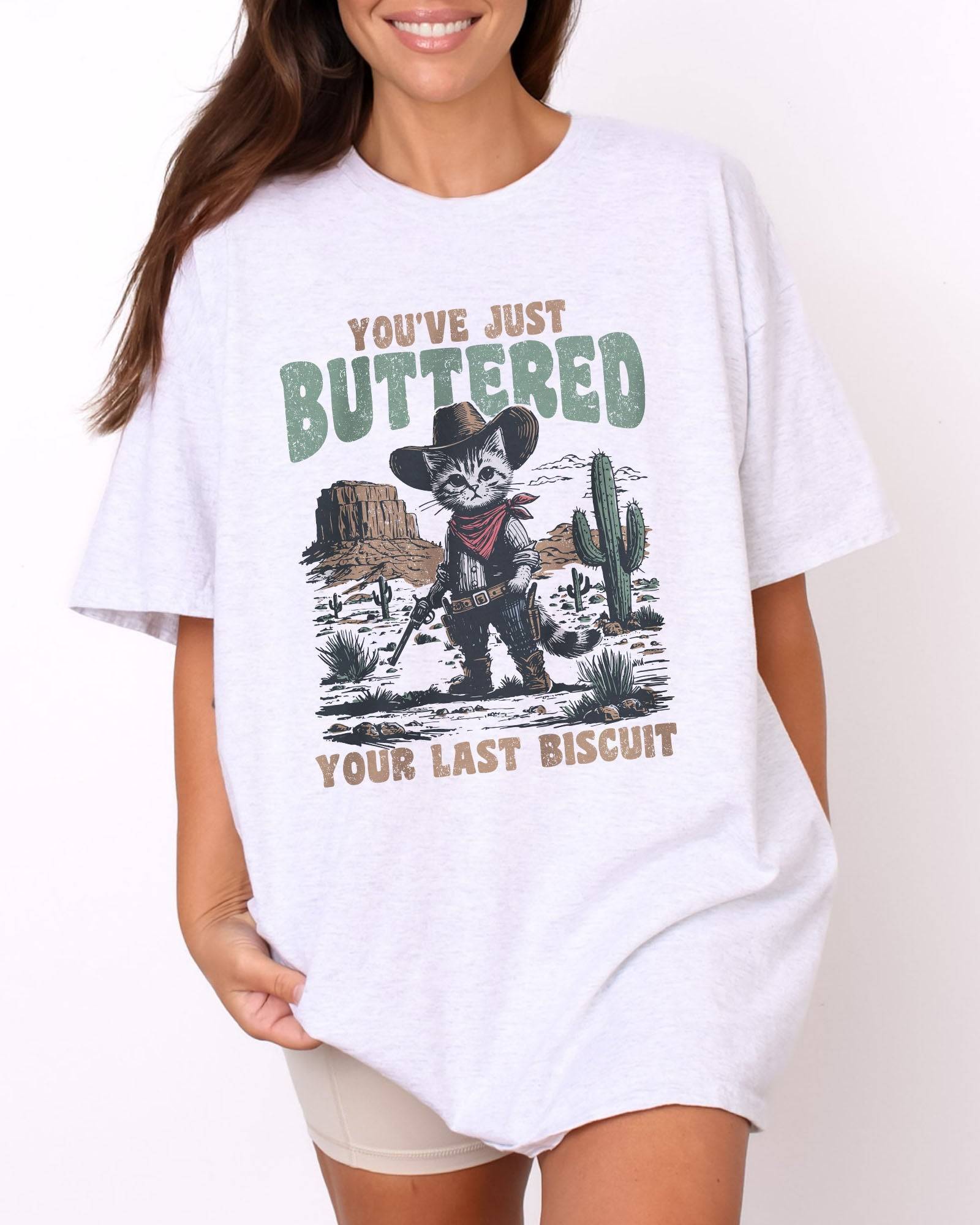 Model wearing ash color shirt with 'You've Just Buttered Your Last Biscuit' cowboy kitten graphic design