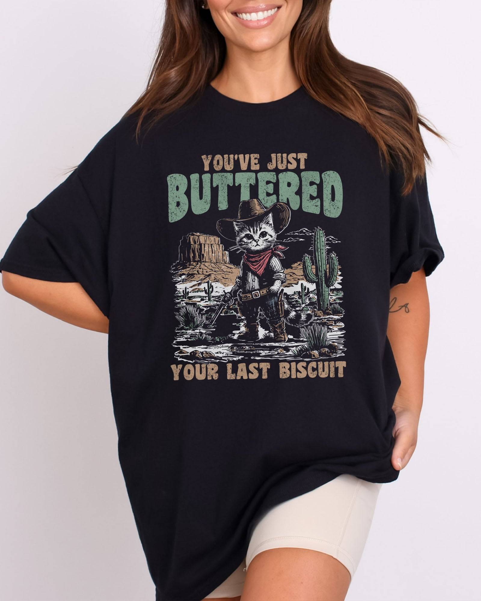 Model wearing black color shirt with 'You've Just Buttered Your Last Biscuit' cowboy kitten graphic design