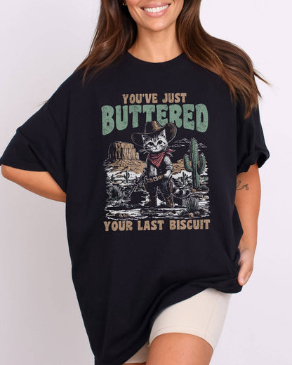 Model wearing black color shirt with 'You've Just Buttered Your Last Biscuit' cowboy kitten graphic design