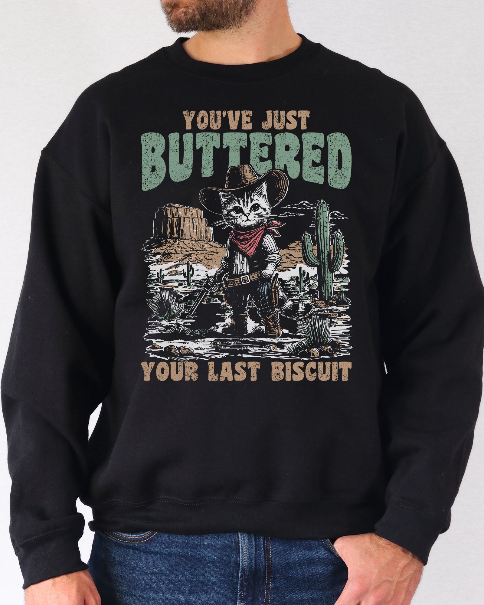 Man wearing a black sweatshirt featuring a funny retro-style cowboy cat graphic and the quote 'You've Just Buttered Your Last Biscuit'.