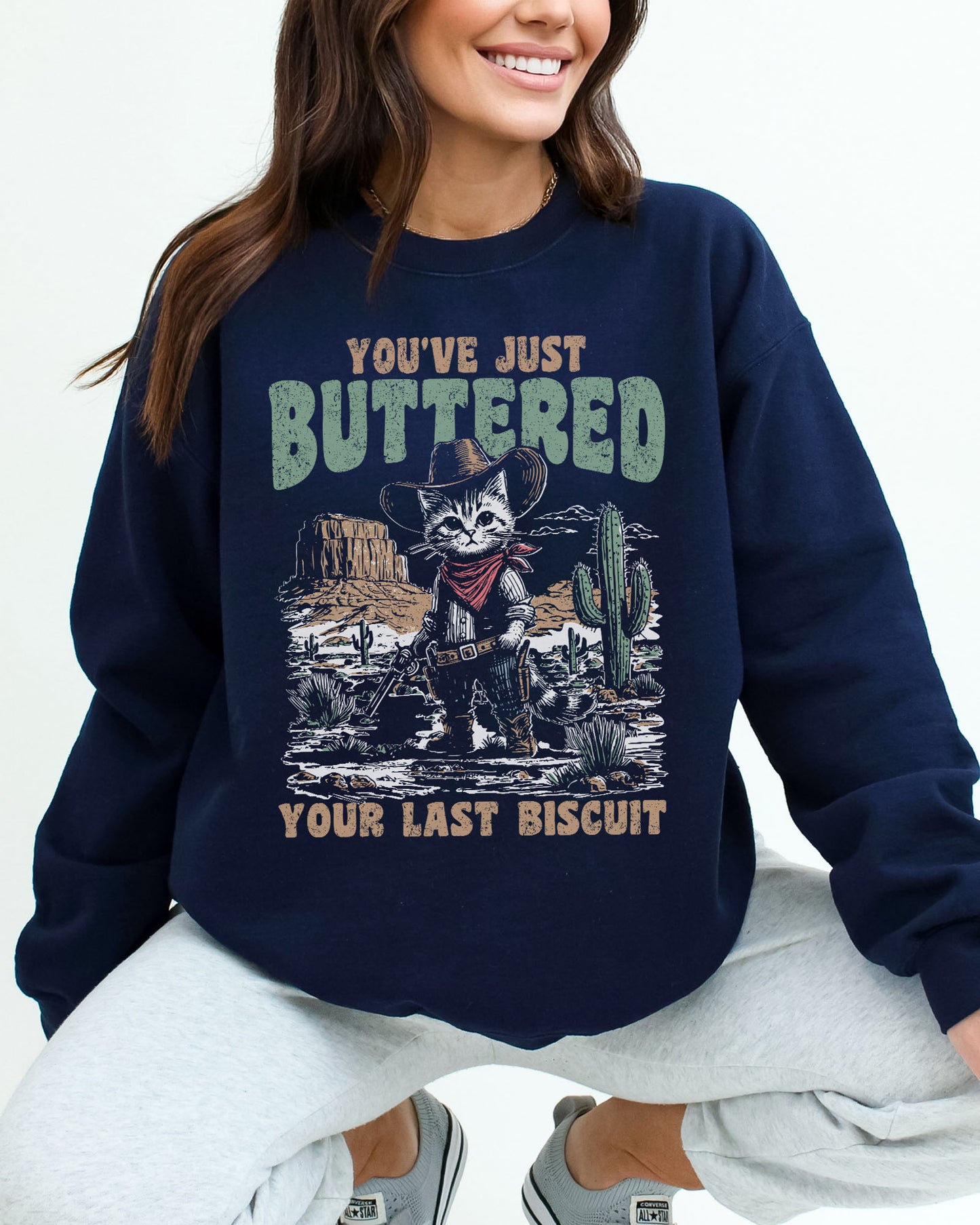 Woman wearing a navy blue sweatshirt featuring a funny retro-style cowboy cat graphic and the quote 'You've Just Buttered Your Last Biscuit'.