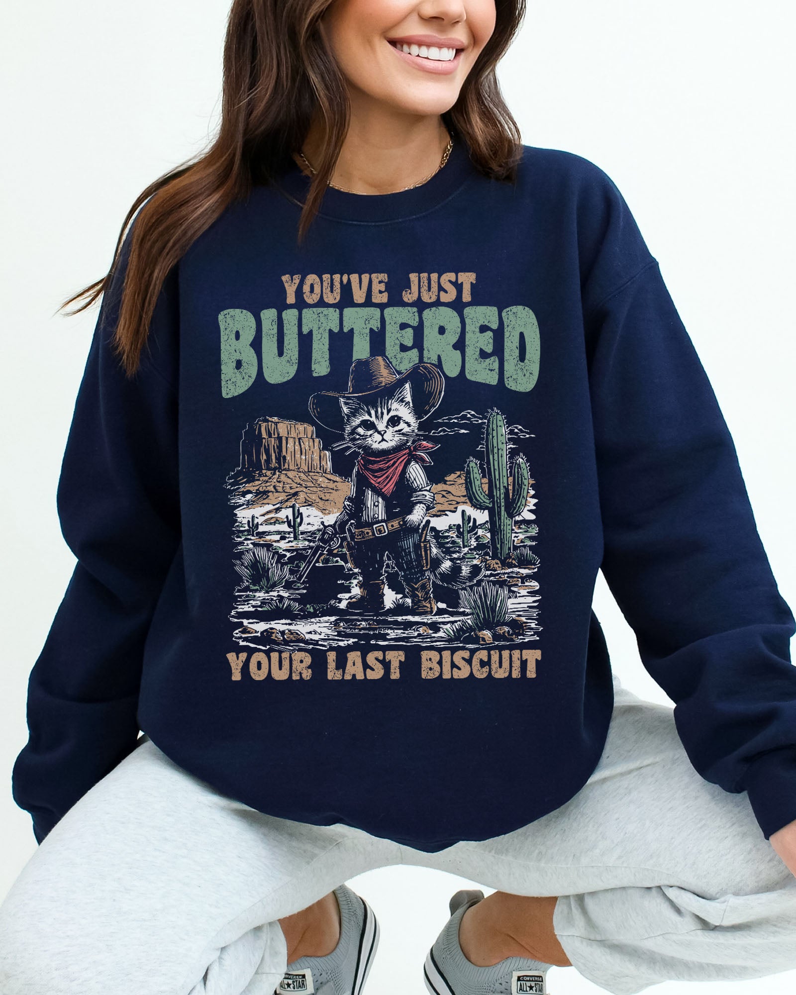 Woman wearing a navy blue sweatshirt featuring a funny retro-style cowboy cat graphic and the quote 'You've Just Buttered Your Last Biscuit'.