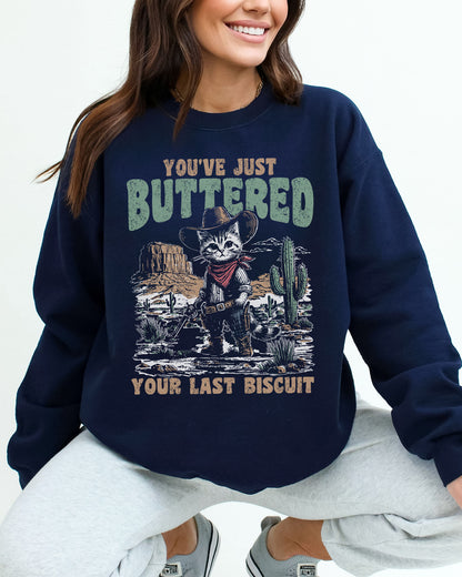 Woman wearing a navy blue sweatshirt featuring a funny retro-style cowboy cat graphic and the quote 'You've Just Buttered Your Last Biscuit'.