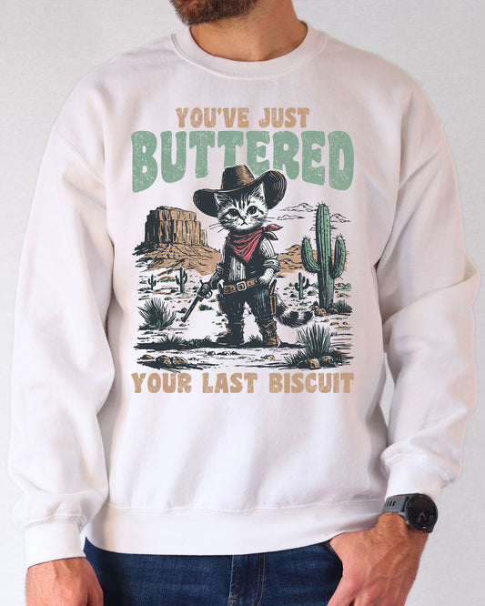 Man wearing a white sweatshirt featuring a funny retro-style cowboy cat graphic and the quote 'You've Just Buttered Your Last Biscuit'.