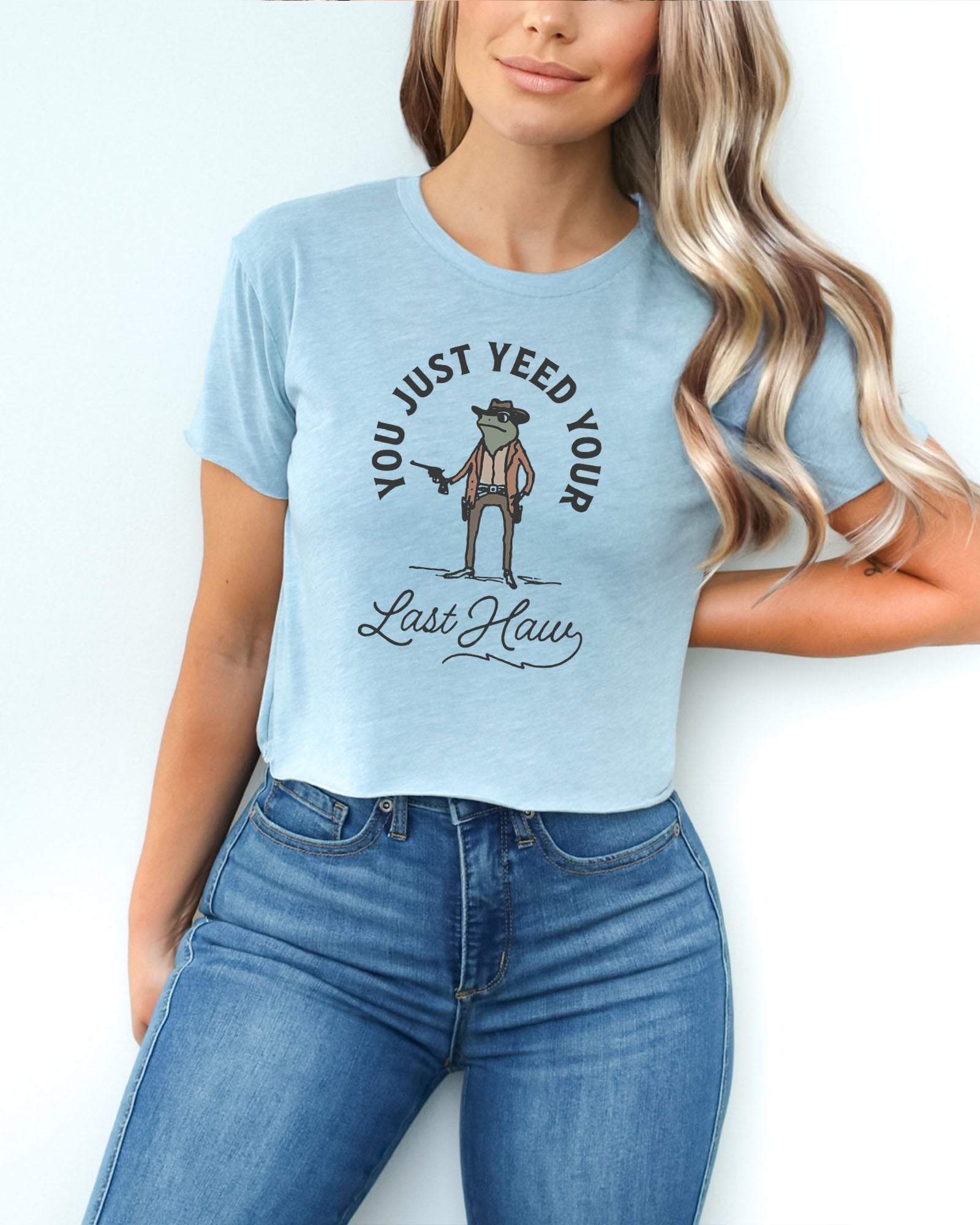 Woman wearing a stonewash denim crop top with a funny retro cowboy sheriff frog graphic and the text 'You Just Yeed Your Last Haw'.