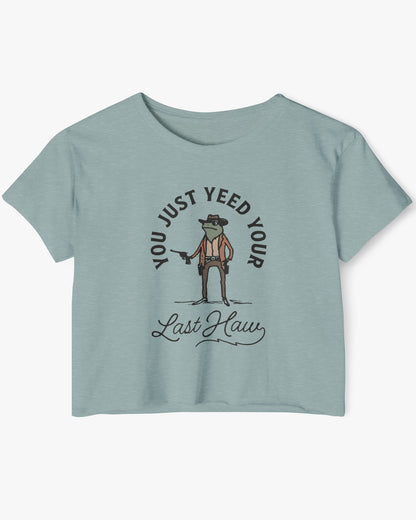 Flat lay of a stonewash green crop top with a funny retro cowboy sheriff frog graphic and the text 'You Just Yeed Your Last Haw'.