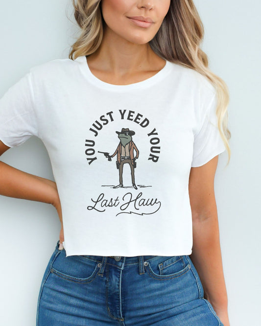 Woman wearing a white crop top with a funny retro cowboy sheriff frog graphic and the text 'You Just Yeed Your Last Haw'.