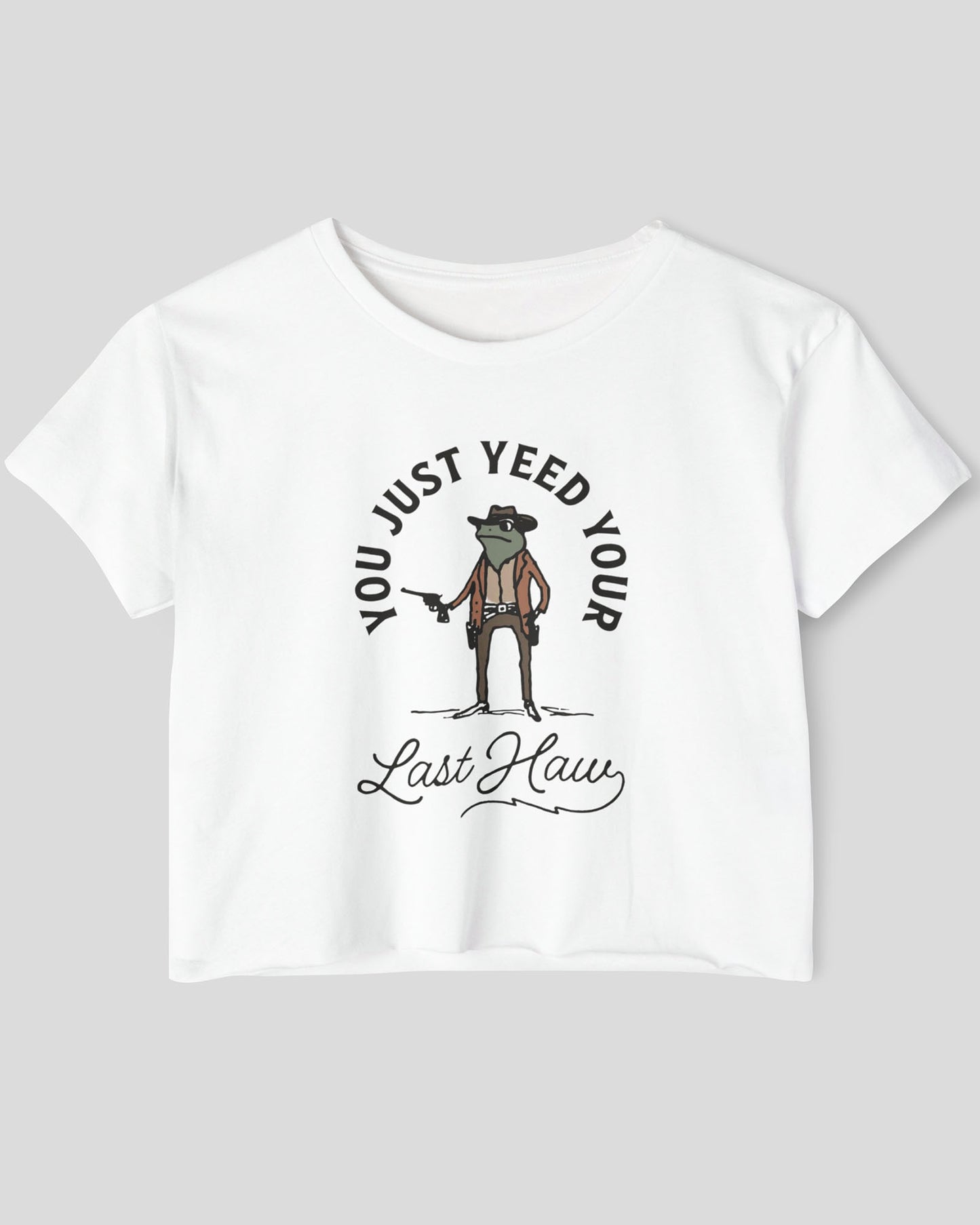 Flat lay of a white crop top with a funny retro cowboy sheriff frog graphic and the text 'You Just Yeed Your Last Haw'.