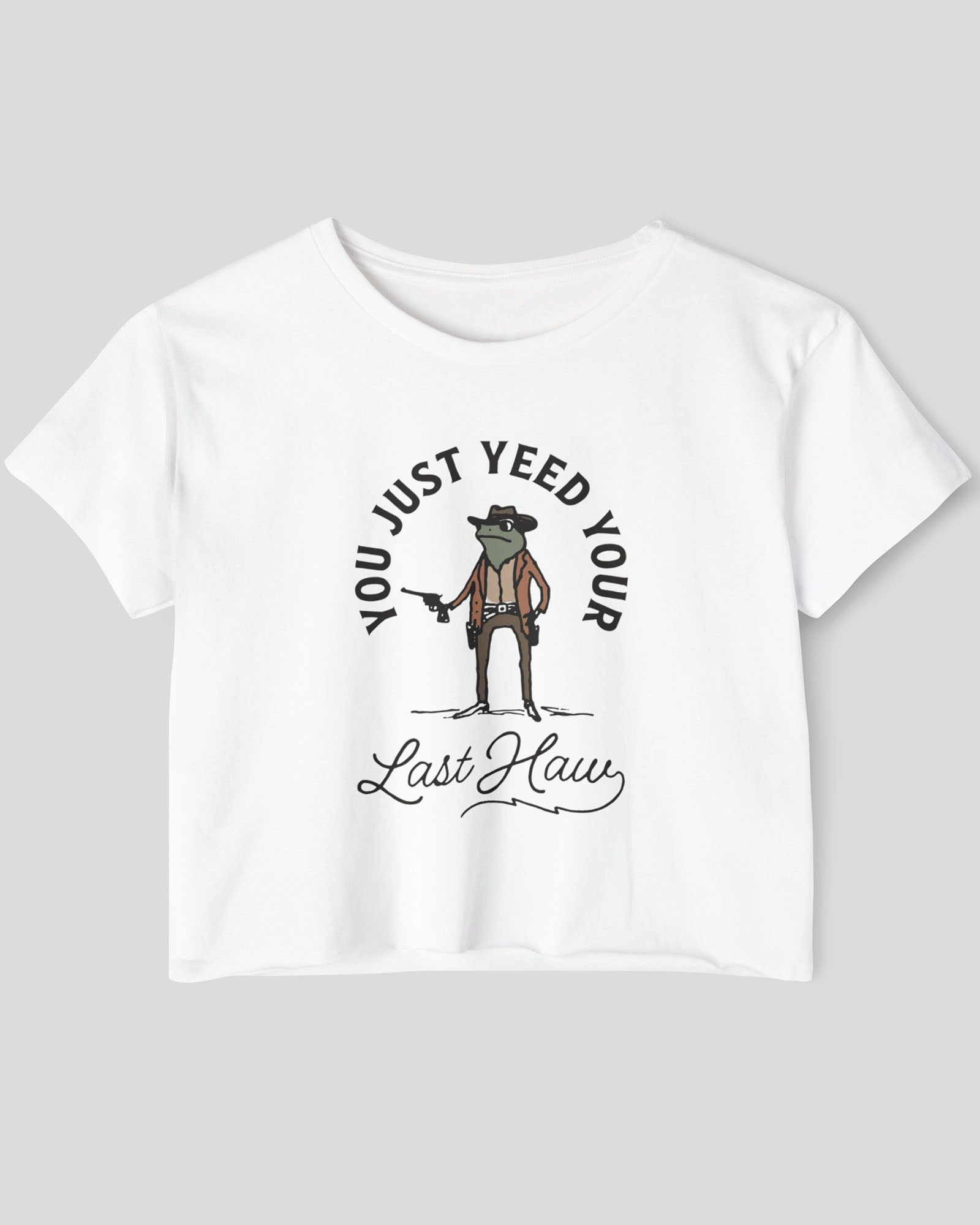 Flat lay of a white crop top with a funny retro cowboy sheriff frog graphic and the text 'You Just Yeed Your Last Haw'.