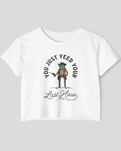 Flat lay of a white crop top with a funny retro cowboy sheriff frog graphic and the text 'You Just Yeed Your Last Haw'.
